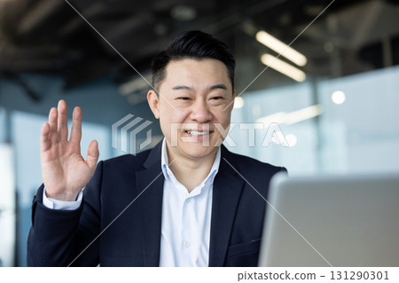 Close-up photo of a young Asian businessman working in an office, sitting at a desk in front of a laptop, talking and greeting the camera. Close-up photo of a young Asian businessman working in an office, sitting at a desk in front of a laptop, talking and greeting the camera. 131290301