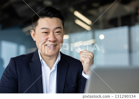 Close-up photo of a young Asian man in a suit sitting in the office in front of a laptop, looking at the screen and happy showing a victory gesture with his hand. Close-up photo of a young Asian man in a suit sitting in the office in front of a laptop, looking at the screen and happy showing a victory gesture with his hand. 131290334