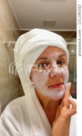 An adult woman in a white coat and a towel on her head applies a cosmetic mask on her face 131290382