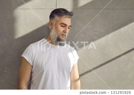 Handsome senior bearded male model in white T shirt standing by gray concrete wall Handsome senior bearded male model in white T shirt standing by gray concrete wall 131291920