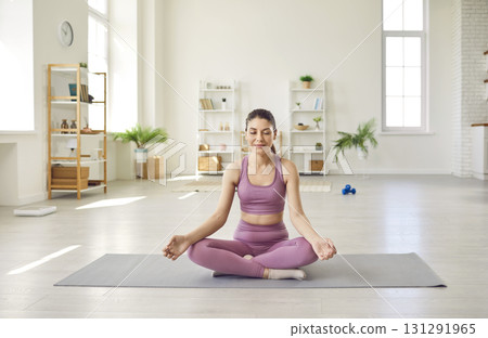 Peaceful sporty young woman doing yoga exercise at home sitting in lotus pose. Peaceful sporty young woman doing yoga exercise at home sitting in lotus pose. 131291965