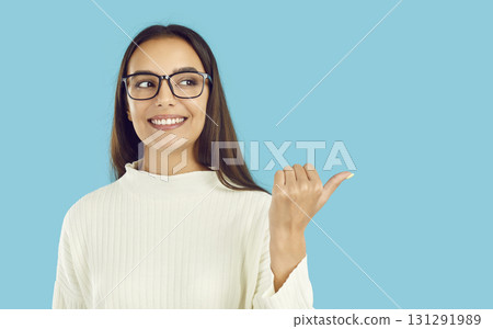 Brunette smiling friendly girl in glasses is pointing thumb on copyspace on blue background. Brunette smiling friendly girl in glasses is pointing thumb on copyspace on blue background. 131291989