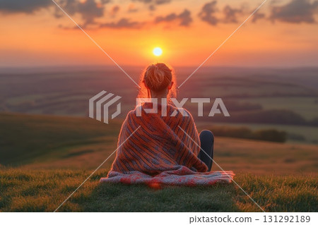 Woman sitting on hill watching warm sunset over valley landscape Woman sitting on hill watching warm sunset over valley landscape 131292189