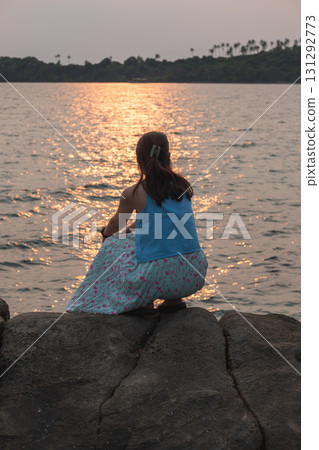 Asian woman sitting on rocks watching sunset over calm sea with distant island Asian woman sitting on rocks watching sunset over calm sea with distant island 131292773