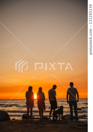 Four people standing with a German Boxer dog on the beach during a stunning sunset, looking at the ocean waves and enjoying a calm evening together. Four people standing with a German Boxer dog on the beach during a stunning sunset, looking at the ocean waves and enjoying a calm evening together. 131293139