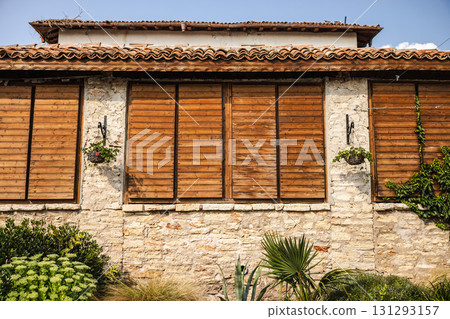 Rustic stone building with closed wooden shutters, tiled roof, and hanging baskets, designed to provide shade and protection from strong summer sun. Rustic stone building with closed wooden shutters, tiled roof, and hanging baskets, designed to provide shade and protection from strong summer sun. 131293157