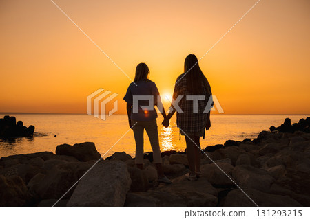 Silhouette of two women holding hands on seaside rocks at sunrise 131293215