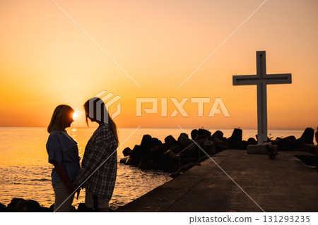 Two women holding hands at sunrise near a large cross monument by the sea in Varna, Bulgaria 131293235