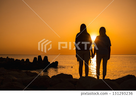 Two women holding hands on the rocks at sunrise, looking at the sea horizon, symbolizing love, unity, support, family bonds, and emotional well-being 131293236