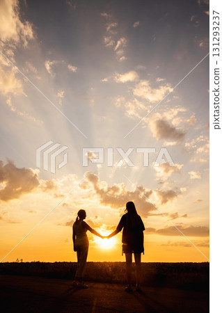 Two women holding hands in silhouette at sunset with dramatic rays of light breaking through the clouds 131293237