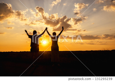 Two women standing in silhouette at sunset with raised arms, celebrating under dramatic sky 131293239
