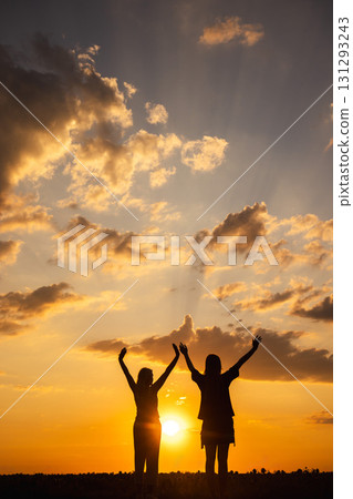 Two women in silhouette raising arms at sunset with dramatic clouds and golden light on the horizon 131293243