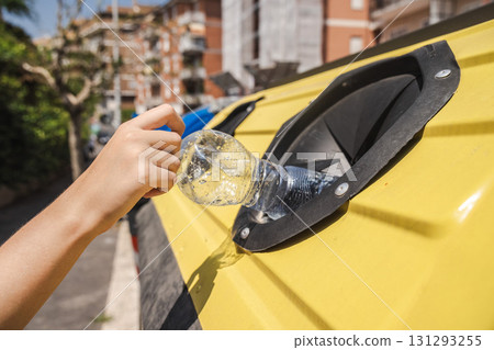Close up of a hand placing a plastic bottle into a yellow recycling bin outdoors under bright sunlight. 131293255