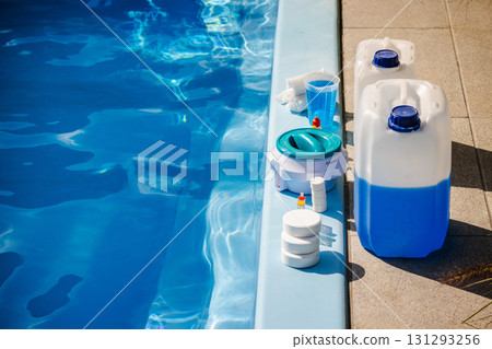 Swimming pool cleaning products including chemical canisters with blue liquid, chlorine tablets, floating dispenser, measuring cup and sanitizer bottles placed at pool edge 131293256