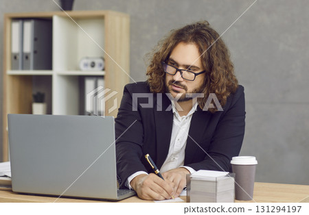 Young businessman sitting at office desk, using laptop computer and taking notes Young businessman sitting at office desk, using laptop computer and taking notes 131294197