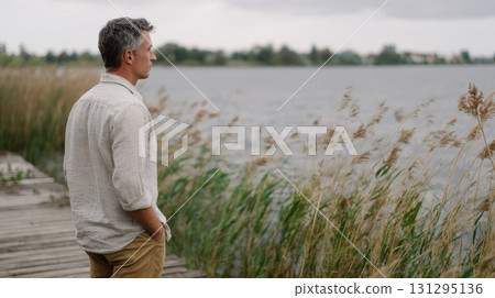 A contemplative man in a linen shirt stands on a peaceful pier gazing at the lake, surrounded by swaying grasses and moody skies, captured in warm vintage film style. 131295136