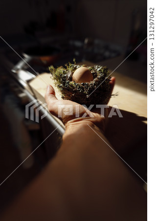A man's hand holding a bird's nest with an egg inside, illuminated by the morning sun. 131297042