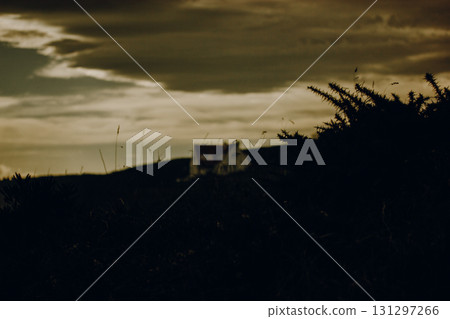 Dark silhouette of a rural house, cottage building on a hill at dusk. A moody landscape features a silhouetted building roof on a hill, with dark foreground and dramatic sky. Rural scene on twilight.  131297266