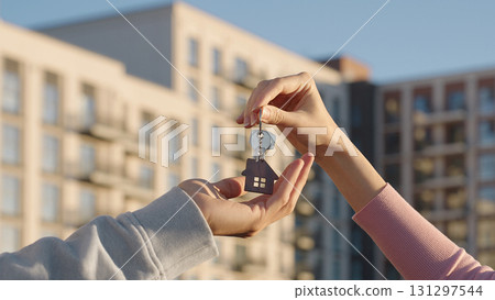 Close up of female real estate owner handing over house key with home shaped keychain to young man outdoors on background of modern apartment building. New home, first time buyer, and property deal. 131297544