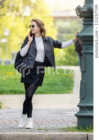Portrait young adult pretty woman wear leather jacket skirt stands outdoors in a park smiling talking smartphone. Stylish confident female person girl enjoy casual street walk sunny day city street 131297599