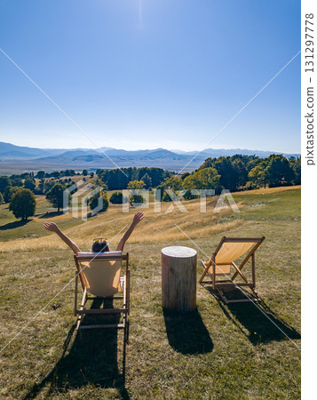 Side view of a woman sitting on a lounge chair with raised arms, overlooking hills and distant mountains in Durmitor Side view of a woman sitting on a lounge chair with raised arms, overlooking hills and distant mountains in Durmitor 131297778
