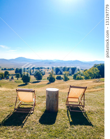 Two lounge chairs with a small table between them, overlooking the hills and distant mountains of Durmitor from a hillside perspective. Two lounge chairs with a small table between them, overlooking the hills and distant mountains of Durmitor from a hillside perspective. 131297779