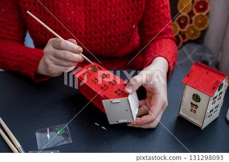 Process of preparing for Christmas and New Year eve. Unrecognizable woman in red sweater painting 3D craft wooden houses DIY. Responsible ecological zero waste handmade decoration. Children hobby 131298093