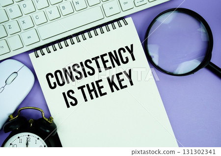 Notepad with the text "Consistency Is The Key" and white keyboard, wireless mouse, magnifying glass and alarm clock 131302341