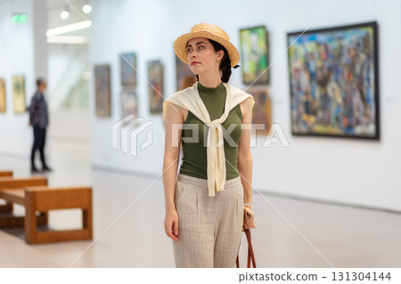 Mid shot of young pretty Caucasian woman in a straw hat and elegant clothes visits a contemporary art gallery. Museum exhibition and cultural education 131304144