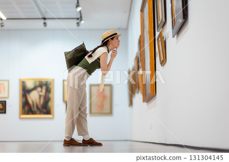 Wide shot of a young Caucasian woman in a straw hat and elegant clothes carefully contemplating modern art in the gallery. Low angle side view 131304145