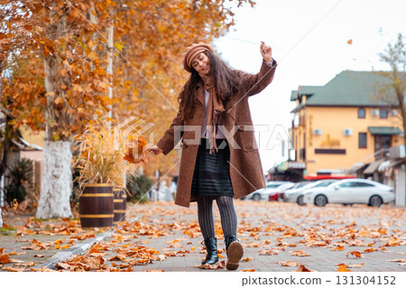 Wrapped in a cozy coat and beret, a Caucasian woman walks dancing through fall streets, savoring her leisure time and the peaceful city environment. 131304152
