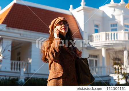 Mid shot of a elegant Caucasian woman in a coat and hat calling by the smartphone and smiling. Beautiful city street in background. Bottom view. Modern communication Mid shot of a elegant Caucasian woman in a coat and hat calling by the smartphone and smiling. Beautiful city street in background. Bottom view. Modern communication 131304161