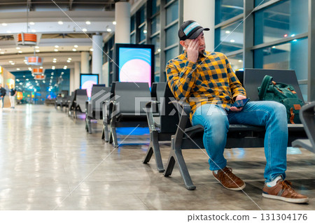 Wide shot of adult Caucasian man in cap and jacket using smartphone in airport waiting area sitting in chair. Low angle view. Concept of travel app Wide shot of adult Caucasian man in cap and jacket using smartphone in airport waiting area sitting in chair. Low angle view. Concept of travel app 131304176