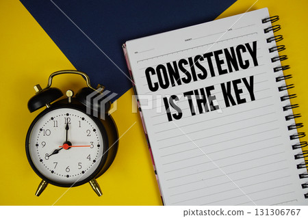 Consistency Is The Key text written on notepad with alarm clock on yellow background 131306767