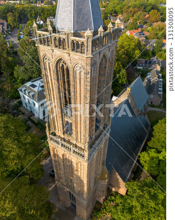 Aerial view of Gothic brick church tower with pointed roof and pinnacles, surrounded by trees and residential houses in a quiet neighborhood. Aerial view of Gothic brick church tower with pointed roof and pinnacles, surrounded by trees and residential houses in a quiet neighborhood. 131308095