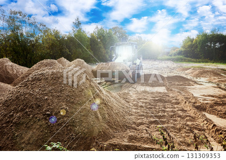 Backhoe loader is engaged in sand distribution work on construction site with several sand piles arranged across working area. Backhoe loader is engaged in sand distribution work on construction site with several sand piles arranged across working area. 131309353