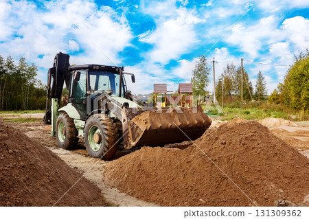 Backhoe loader stands on site with bucket facing large sand mound, surrounding area covered with multiple piles prepared for construction activity. Backhoe loader stands on site with bucket facing large sand mound, surrounding area covered with multiple piles prepared for construction activity. 131309362