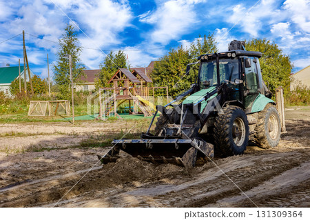 Loader moves sand in front of wooden playground structure, small houses and trees visible in background of work site. Loader moves sand in front of wooden playground structure, small houses and trees visible in background of work site. 131309364