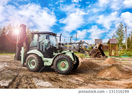 Backhoe loader operates on construction ground with several sand piles placed nearby as site undergoes preparation for further work. 131310299