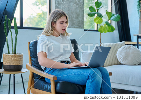Portrait of young woman sitting in chair typing on laptop, in home interior Portrait of young woman sitting in chair typing on laptop, in home interior 131311099