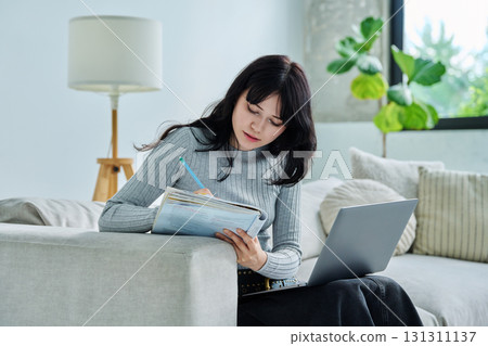 Young student girl study online, sitting at home on couch with laptop, textbook Young student girl study online, sitting at home on couch with laptop, textbook 131311137