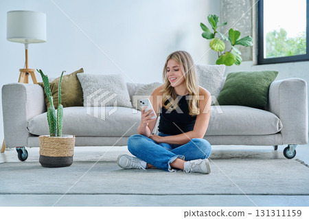 Smiling young woman with smartphone sitting at home near sofa Smiling young woman with smartphone sitting at home near sofa 131311159