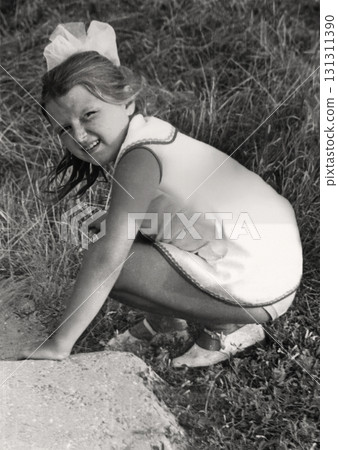 A vintage portrait of a seven-year-old girl in a short dress with a white bow in her hair, crouched and searching for something in the grass. Photo from 1974. 131311390