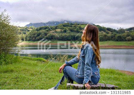 Young woman sitting on rock by lake looking at mountains Young woman sitting on rock by lake looking at mountains 131311985