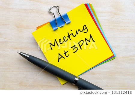 Meeting at 3PM handwritten text on yellow notepad with silver clip top view on wooden table. 131312418