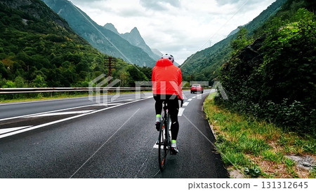 Follow camera view of a cyclist in red jacket riding on the road to Oz Lake in the French Alps, surrounded by dramatic green mountains and cloudy sky, symbolizing endurance and freedom 131312645