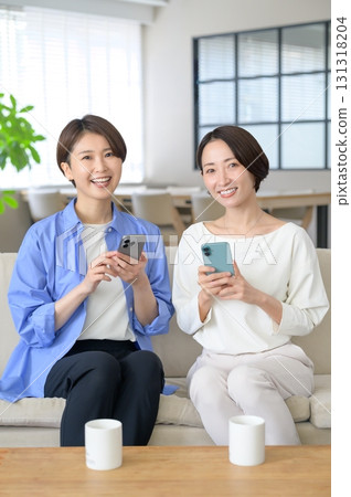 女人們並排坐在沙發上，看著自己的智慧型手機 131318204