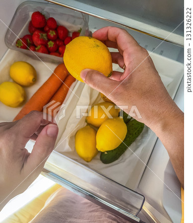 Close-up of hand holding a lemon over fresh vegetables and fruits in a fridge drawer. Healthy lifestyle and nutrition concept. High quality photo 131326222
