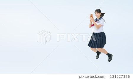 A full-body photo of a female student wearing a sailor suit jumping while looking at her smartphone 131327326