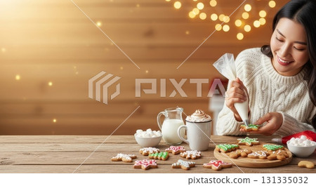 Young Asian woman decorating festive Christmas cookies with white icing using piping bag at wooden table with hot cocoa and fairy lights in background 131335032